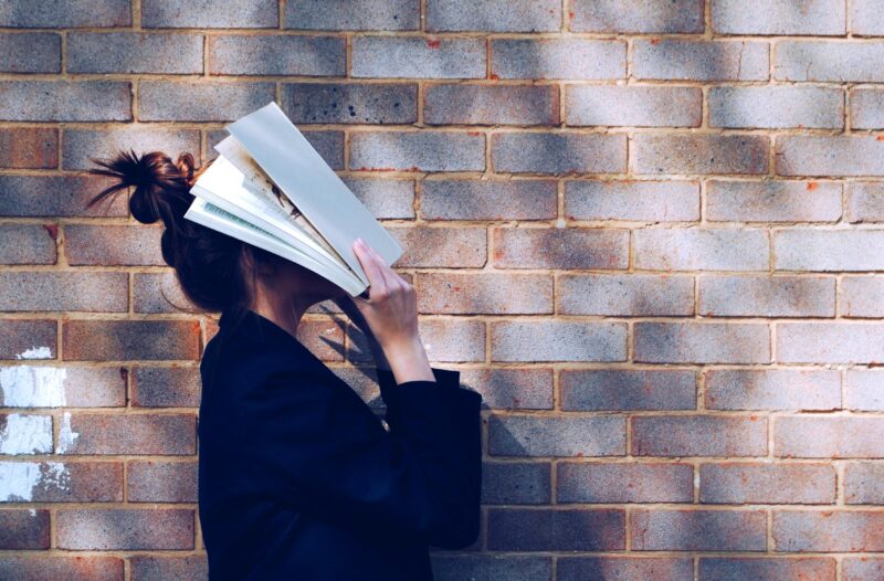Student leaning against brick wall with an open book covering their face