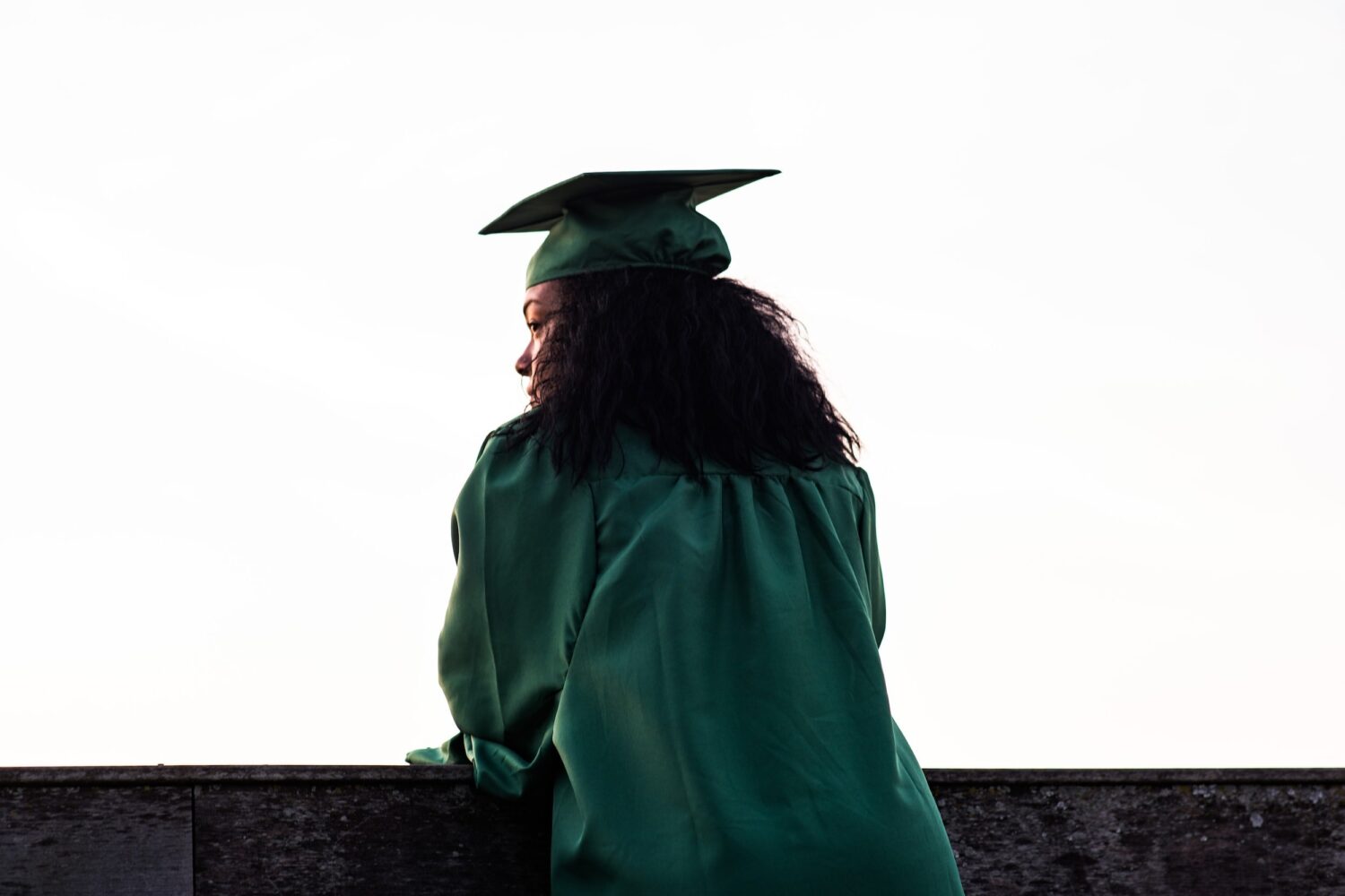 Graduate in green cap and gown standing outdoors looking into the distance