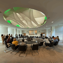 People seated in a round conference room with a large ceiling design and projector.