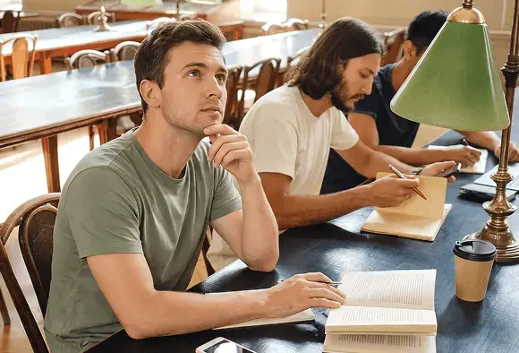 Three men seated at a table in a library, engaged in discussion with books and papers around them.