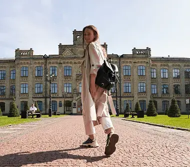 A woman walks along a brick path in front of a building, surrounded by greenery and sunlight.
