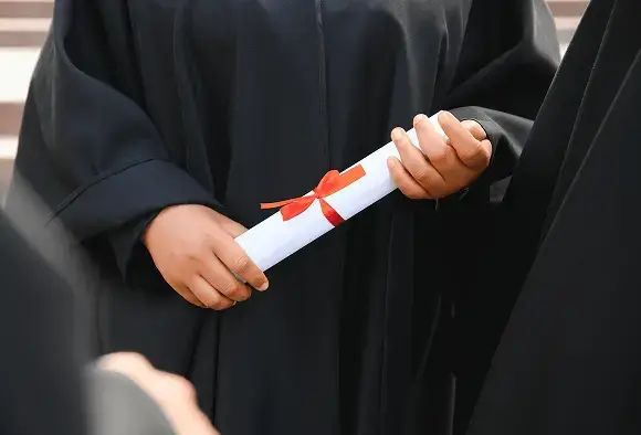 A graduate in a black gown proudly holds a diploma, smiling at the camera in a celebratory pose.