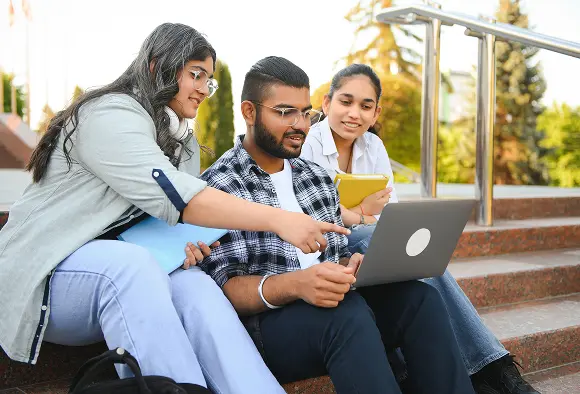 Three students seated on steps, collaborating on a laptop, engaged in discussion and sharing ideas.
