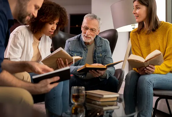 A diverse group of individuals seated at a table, engaged in reading various books together.