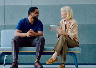 A man and woman sit on a bench, both focused on a tablet in their hands, sharing a moment of engagement.