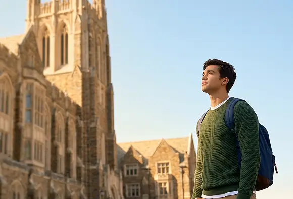 A man with a backpack stands in front of a large building, looking towards the entrance.