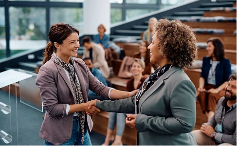 Two women shake hands in front of a diverse crowd, symbolizing collaboration and unity.