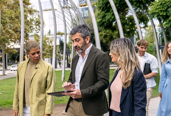 Several individuals gather in a park, focused on a tablet, with lush grass and trees in the background.