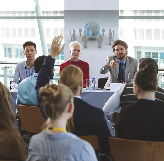 A group of people seated at a table, with one individual raising their hand to speak or ask a question.