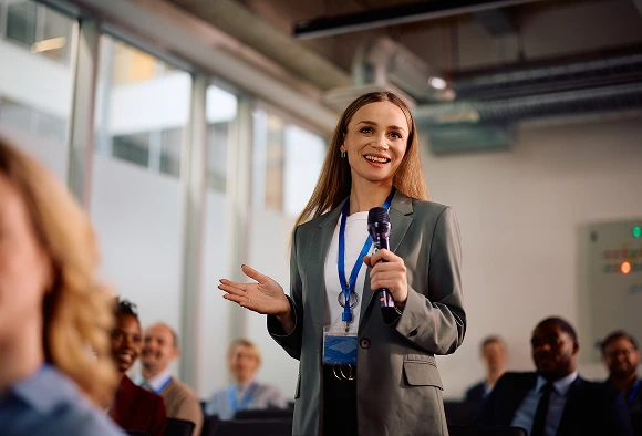 A woman presents to an audience, gesturing confidently while discussing her topic.