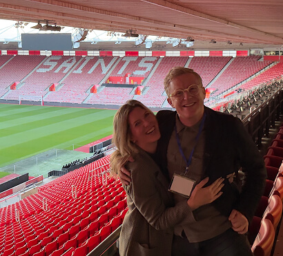 A man and a woman pose for a photo in a stadium, showcasing the seating and field behind them.