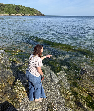 A woman stands on the shore, gazing thoughtfully at the water under a clear sky.