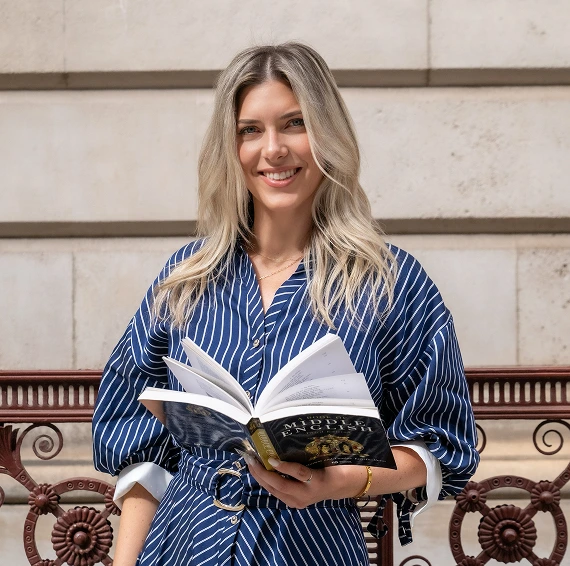 Jackie in a blue and white striped dress holds a book, smiling as she stands outdoors.