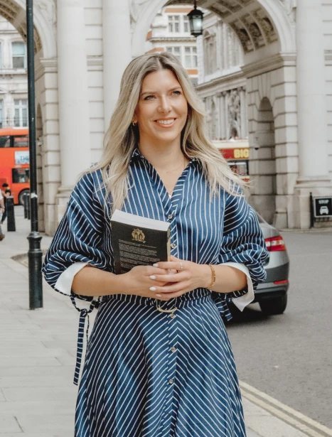 Jackie in a blue and white striped dress is holding a book, standing in a relaxed pose outdoors.