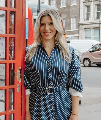 Jackie in a blue and white striped dress stands beside a red telephone booth, smiling at the camera.