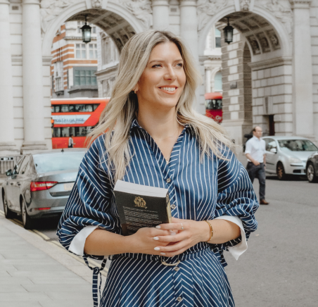 Jackie in a blue and white striped dress is holding a book, smiling while standing outdoors.