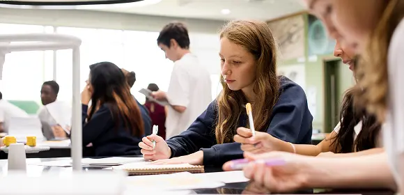 A group of students seated at desks in a classroom, focused on writing in their notebooks.