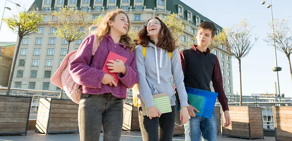 Three young individuals stand together on a sidewalk, smiling and engaged in conversation.