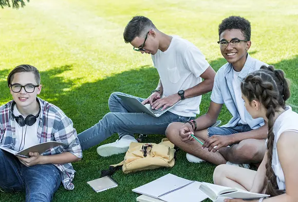 Four young individuals seated on grass, each using a laptop, engaged in a collaborative work or study session.