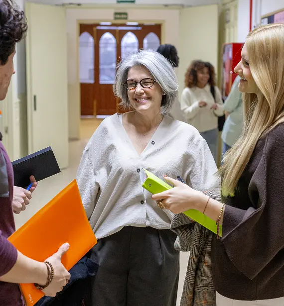 Three women engaged in conversation in a hallway, with one woman gesturing while the others listen attentively.