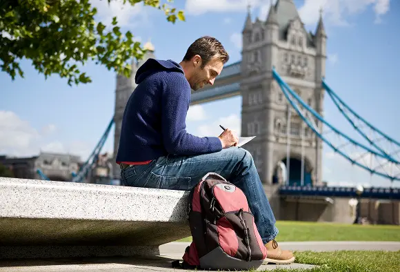A student sits at a table, writing notes with Tower Bridge in the background, showcasing a blend of study and iconic architecture.